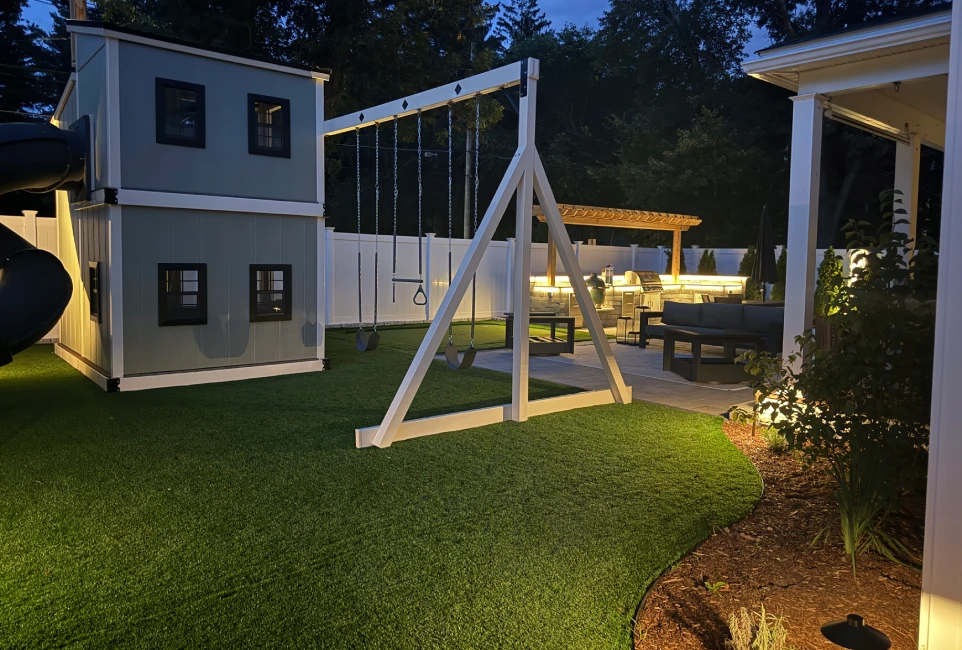 Two-story gray play cottage with spiral slide at dusk, lit by landscape lights, Newton MA