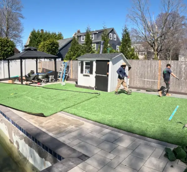 Crew installing artificial turf and gray paver pool deck at Lexington Massachusetts backyard with pool and pergola